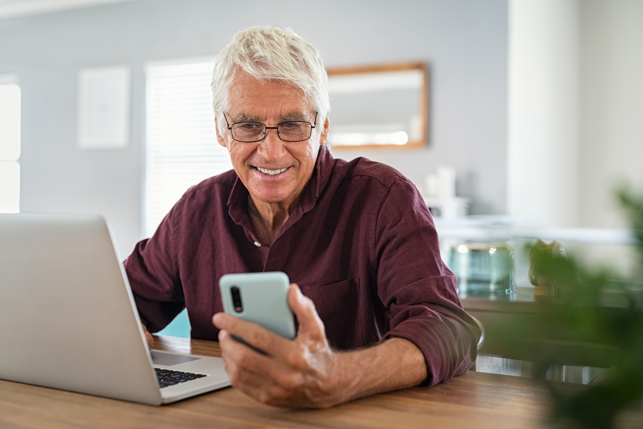 older man with glasses smiling at phone with laptop in front of him