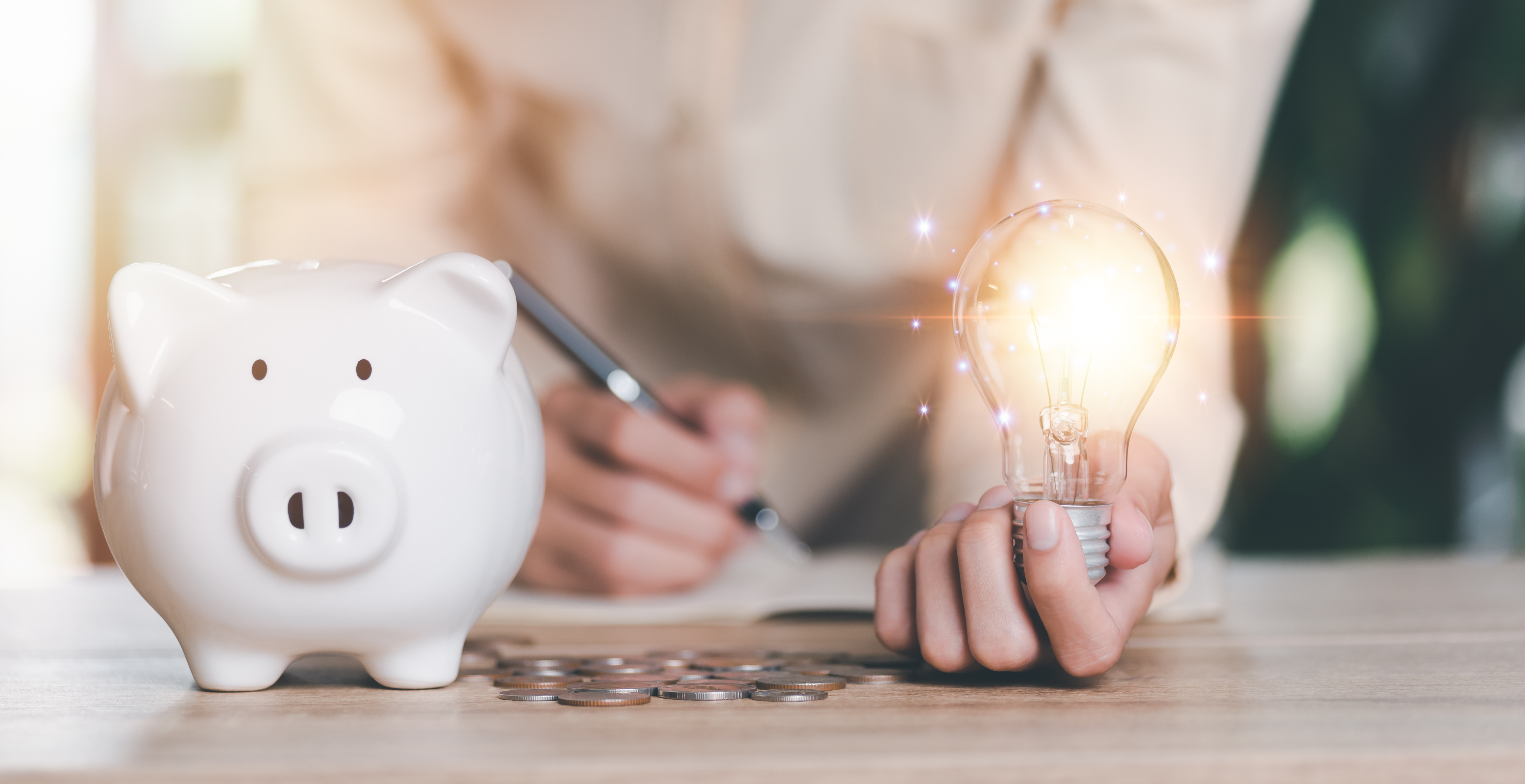 white piggy bank on table with hand holding lit up light bulb