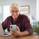 older man with glasses smiling at phone with laptop in front of him