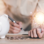 white piggy bank on table with hand holding lit up light bulb